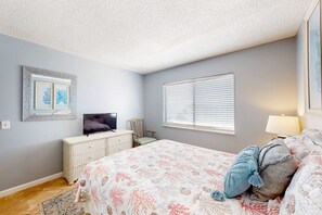 Ein Schlafzimmer mit einem Bett, einem Schminktisch, einem Fernseher, einem Fenster mit Jalousien und einem Bild in einem Rahmen an der Wand.