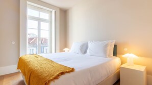 A bedroom with a large bed, white bedding, a mustard-colored throw blanket, a nightstand with a lamp, and a window with a view of buildings. - LovelyStay - Entreparedes House - 302 (Porto)