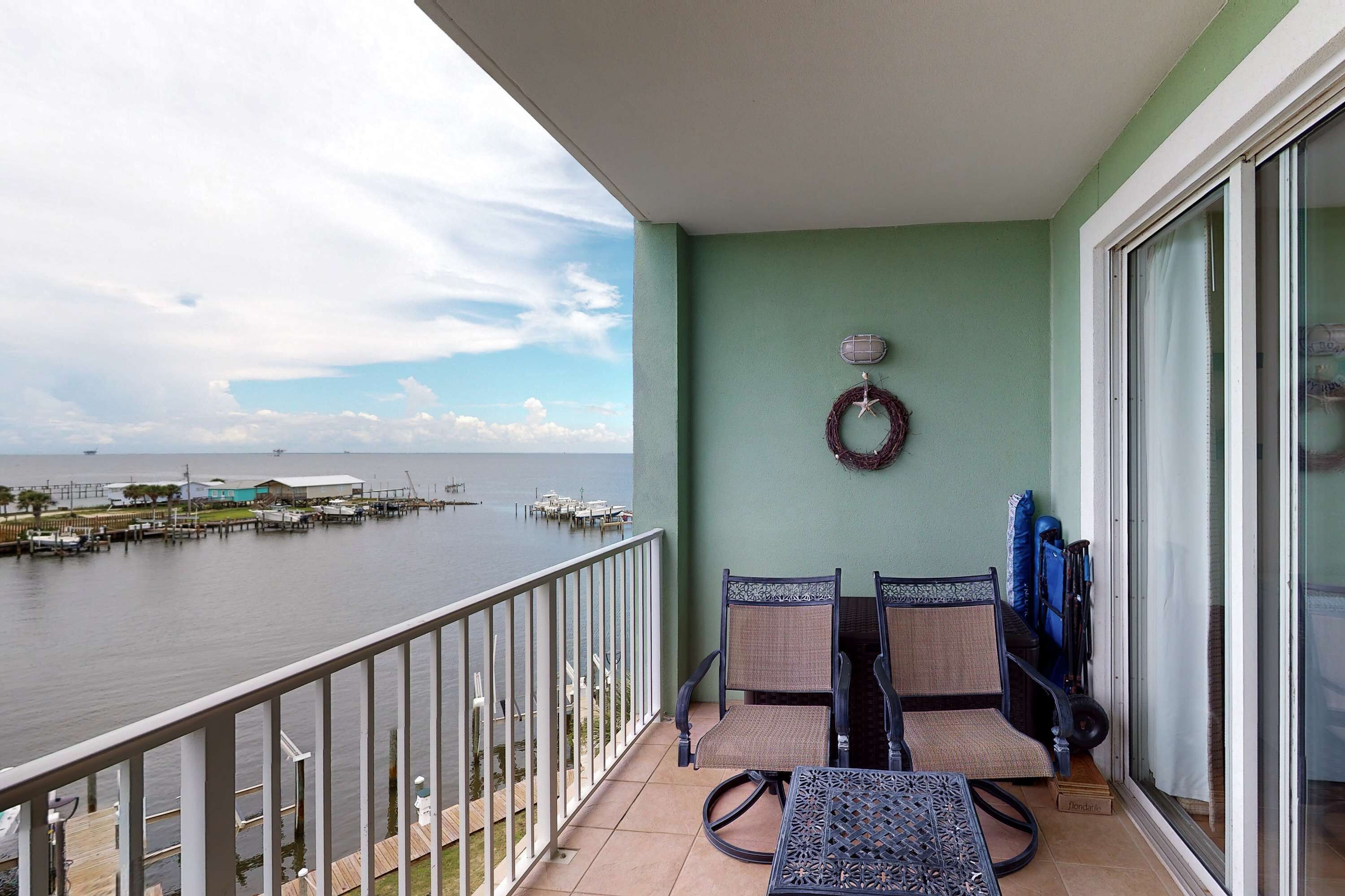 A balcony with a view of the water, a table, two chairs, and a wreath on the wall.