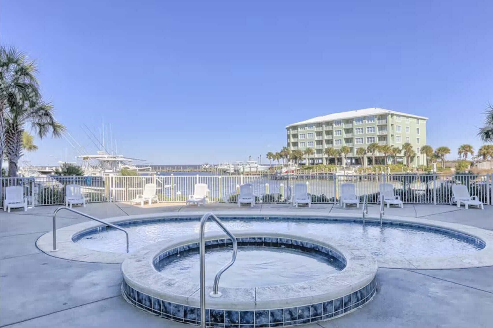 A hot tub with a circular design, surrounded by lounge chairs, with a marina and a hotel in the background.