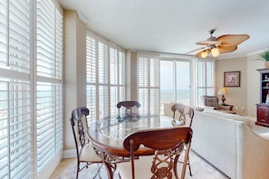 A dining area with a glass-top table, wooden chairs, and a ceiling fan. - Gulf-view condo with direct beach access, indoor & outdoor pools, hot tub, sauna (Gulf Shores)