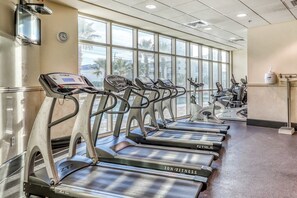Une salle de sport équipée de tapis de course, de vélos d’appartement et dotée d’une grande fenêtre offrant une vue sur des palmiers.