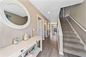 A hallway with a staircase, a round mirror, a console table with decorative items, and a light-colored wall. - Remodeled, lagoon-front home with decks, views, paddleboat (Gulf Shores)