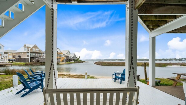 A wooden deck with blue Adirondack chairs and a swing, overlooking a body of water with buildings in the background. - Remodeled, lagoon-front home with decks, views, paddleboat (Gulf Shores)