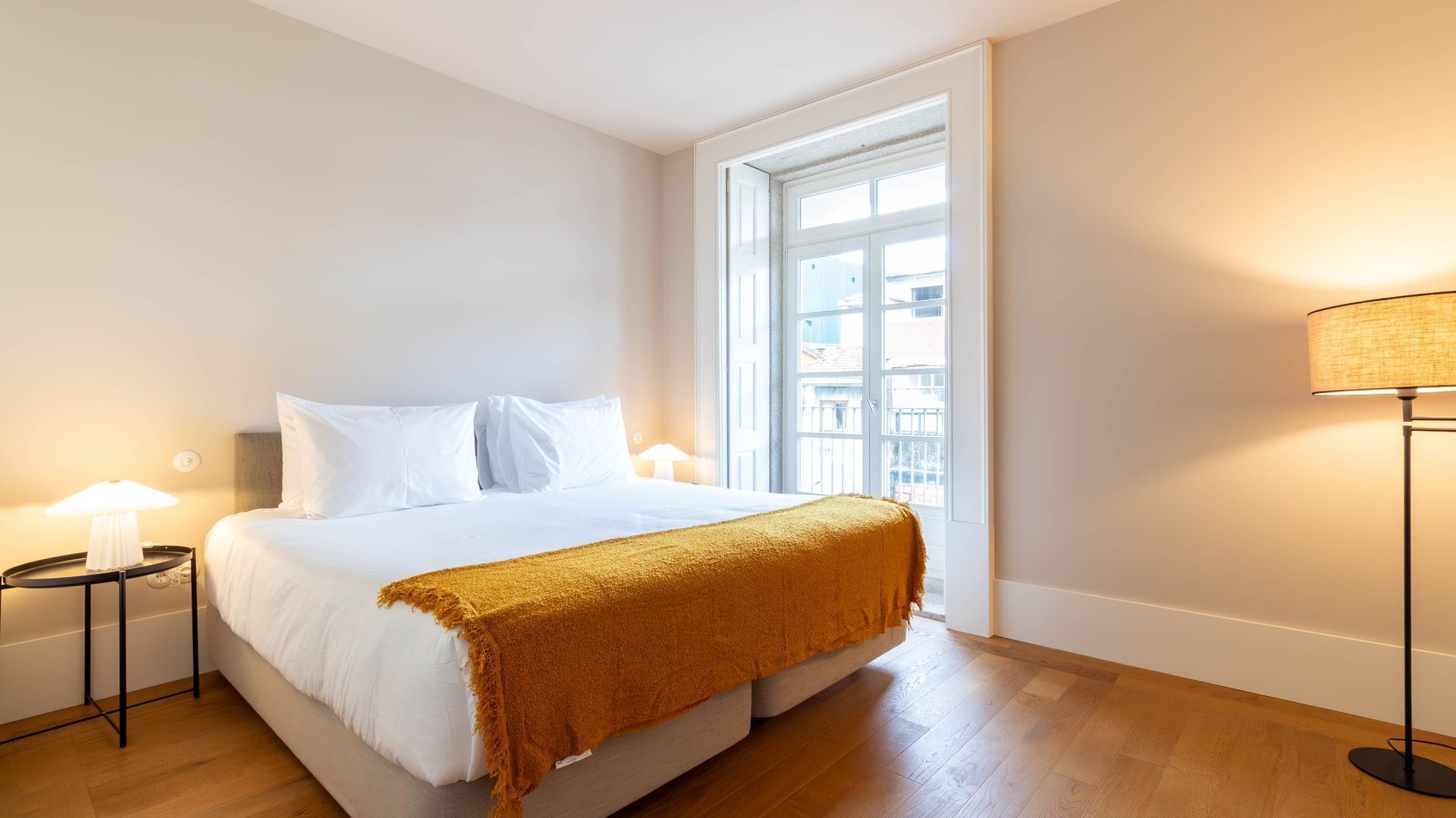 Une chambre d’hôtel moderne avec un grand lit, une fenêtre donnant sur des immeubles et une lampe sur pied.