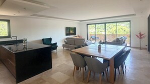 A modern kitchen with a central island, a dining area with a wooden table and chairs, a sofa, and a large window with a view of greenery. - Villa and pool Pacha Prestige (Roquebrune-sur-Argens)