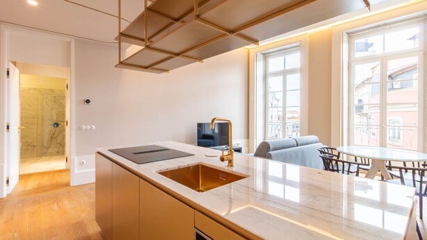 Cozinha moderna com bancada de mármore, torneira dourada e forno embutido. Possui área de estar com mesa e cadeiras, e uma grande janela que oferece vista da cidade.