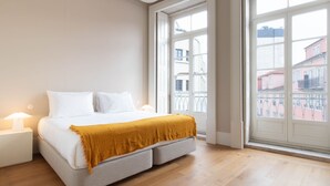 Quarto com cama, luminárias de cabeceira, janela com vista para edifícios e varanda.