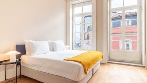 Ein Hotelzimmer mit einem großen Bett, einem Nachttisch mit Lampe und einem Fenster mit Blick auf Gebäude.