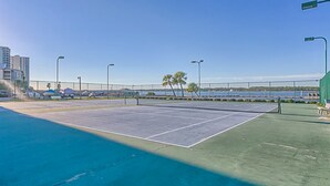 An outdoor tennis court with a net, surrounded by a fence, and a view of a body of water and buildings in the background.