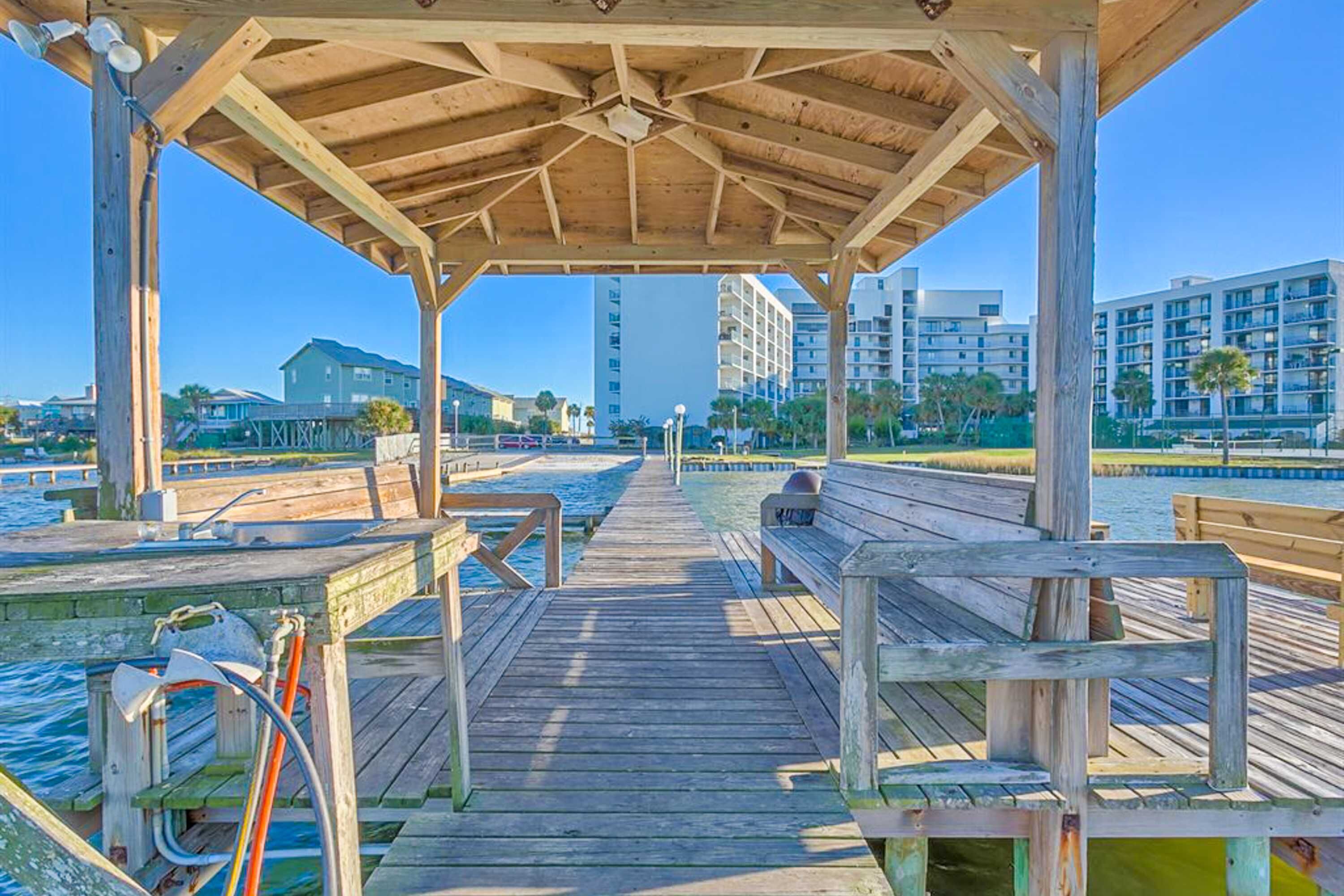 A wooden pier leading to a covered area with benches, overlooking a body of water and buildings in the background.