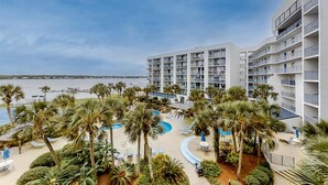 A hotel with multiple floors, a pool area with palm trees, and a view of the beach.