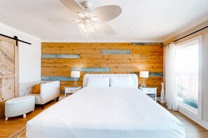 A bedroom with a large bed, two bedside tables with lamps, a ceiling fan, wooden wall paneling, and a window with curtains. - Gulf-Front Condo w Balcony, Beach Views, WiFi, AC & Shared Gulf-Facing Pool (Gulf Shores)
