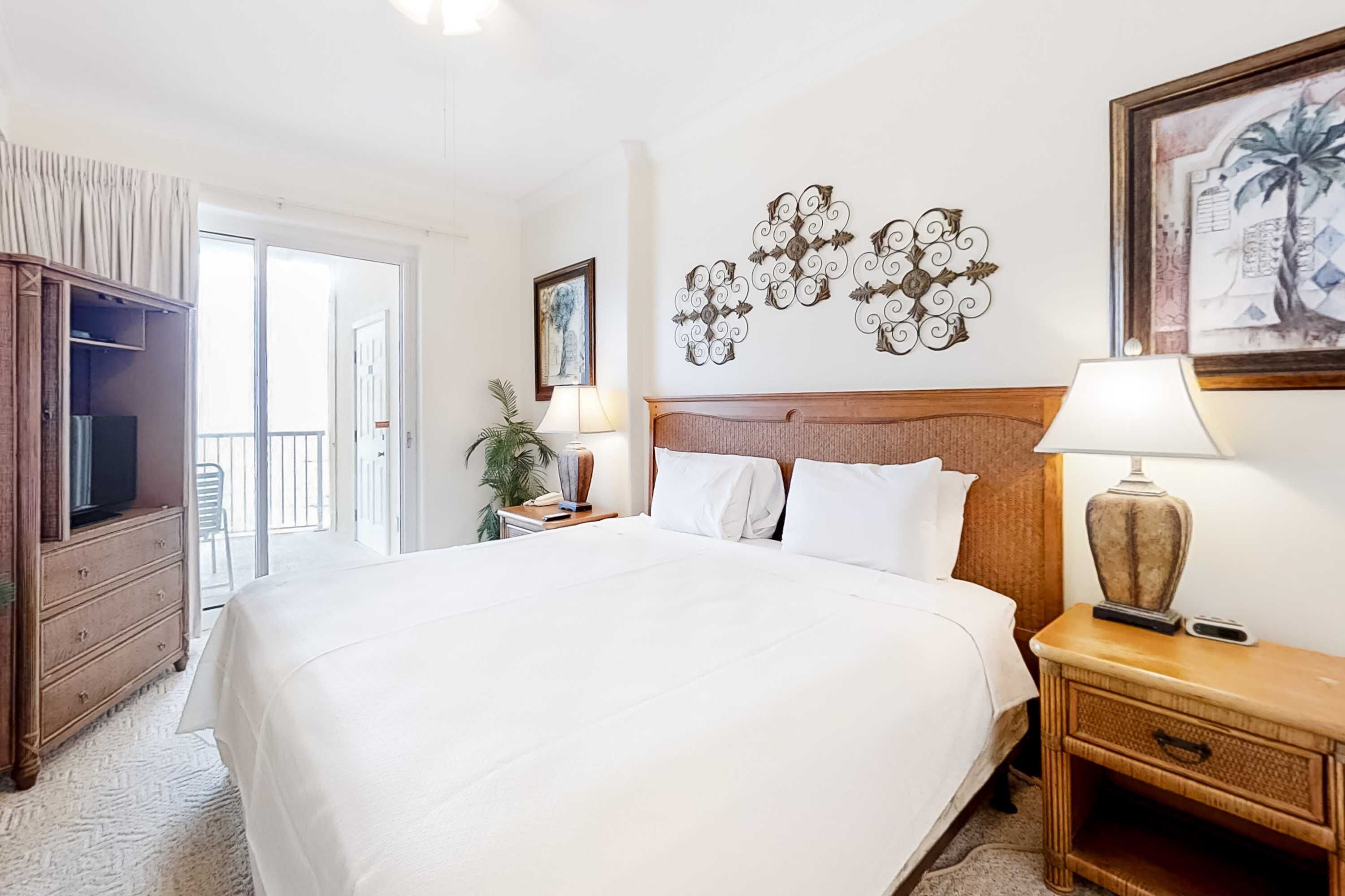 Ein Hotelzimmer mit einem großen Bett, einem Holz-Kopfteil, zwei Nachttischen mit Lampen, einer Kommode aus Holz, einem großen Fenster und Wandbildern.