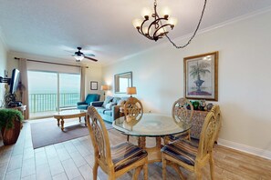A living room with a glass-top dining table, wooden chairs, a ceiling fan, a chandelier, a sofa, and a large window with a view. - Family-Friendly Gulf-Front Condo w/Shared Pool, Hot Tub, Sauna, Private Balcony (Gulf Shores)