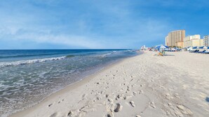 En strand med fotspår i sanden, en klar himmel och byggnader i bakgrunden.