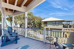 A covered porch with Adirondack chairs, a small round table, and a view of a green building across the street. - Bright home with decks, WiFi, AC, laundry & shared pool dock - walk to the beach (Gulf Shores)