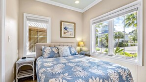 Un dormitorio con cama, mesita de noche, lámpara, un cuadro enmarcado y ventanas amplias.