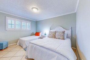 A bedroom with two beds, a window with blinds, a bedside table with a lamp, and a blue stool. - Cheerful beach stay with heated pool, balcony, gulf views, W&D, & central AC (Gulf Shores)