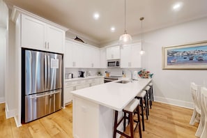 A modern kitchen with white cabinetry, stainless steel appliances, a central island with bar stools, and a coastal-themed painting on the wall. - New home near the beach with on-site pool,hot tub, private balcony, & W&D (Gulf Shores)