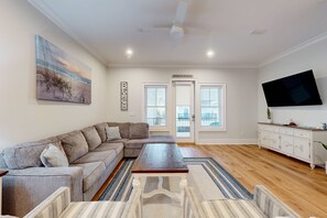 A living room with a sectional sofa, a coffee table, a flat-screen TV, and a painting on the wall. - New home near the beach with on-site pool,hot tub, private balcony, & W&D (Gulf Shores)