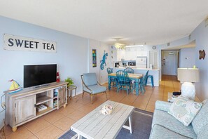 A living room with a television, a coffee table, a sofa, and a dining area with chairs and a table. - Newly updated beachfront condo with Gulf view, indoor,outdoor pools & hot tub (Gulf Shores)