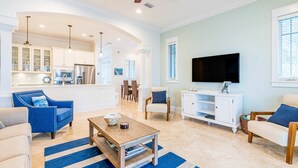 Un salon spacieux avec une cuisine à l’arrière-plan, un téléviseur à écran plat et un ventilateur de plafond.
