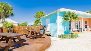 Une terrasse en bois avec des tables de pique-nique, un espace aménagé au bord de la piscine et un bâtiment à la façade bleue.