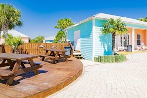 A wooden deck with picnic tables, a poolside area, and a building with a blue exterior. - Modern beach home with screened porch, on-site pool, AC, & W&D (Gulf Shores)
