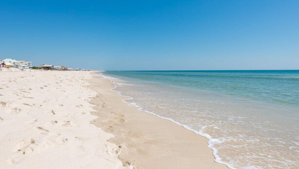 Une plage avec un ciel dégagé, un rivage sablonneux et des bâtiments en arrière-plan.