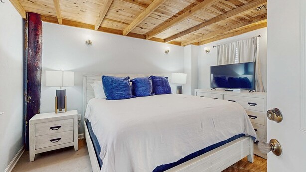 A bedroom with a wooden ceiling, a bed with a white and blue bedspread, two bedside tables with lamps, a television, and a door with a keyhole. - Downstairs duplex near the beach with full kitchen, WiFi, cable, partial AC (Gulf Shores)