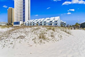 A beachfront hotel with a modern design, featuring a prominent white building with multiple balconies, situated on a sandy beach with sparse vegetation. - First-floor dog-friendly condo with on-site pool, beach access, & balcony (Gulf Shores)