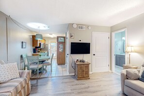 A living room with a sofa, a dining table with chairs, a TV mounted on the wall, and a small cabinet. - First-floor dog-friendly condo with on-site pool, beach access, & balcony (Gulf Shores)