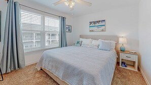 Une chambre à coucher avec un lit, une table de chevet, un ventilateur de plafond, une fenêtre avec des rideaux et un tableau au mur.