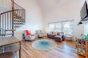 A spacious living room with a wooden floor, a round blue rug, a grey sofa, a small round table, and a staircase with a metal railing. - Sunlit, two-story condo with pool, walk one block to the beach (Gulf Shores)