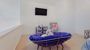 Une pièce comprenant un fauteuil bleu rembourré, un téléviseur fixé au mur, une petite table avec des boîtes de rangement et un fauteuil agrémenté d’un coussin décoratif.