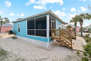 A single-story house with a covered porch and a wooden staircase leading to a deck. - Brand-new cottage with covered patio, central AC, W&D, & pool access - near beach (Gulf Shores)