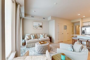 A living room with a sofa, ottoman, and a dining area with a bar stool. - Brand-new beach condo with pool, hot tub, AC & washer/dryer (Gulf Shores)