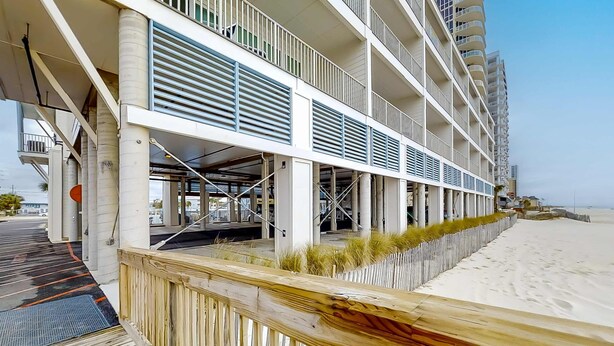 Un hotel de varios pisos con balcones, un área exterior cubierta y una playa de arena al fondo.