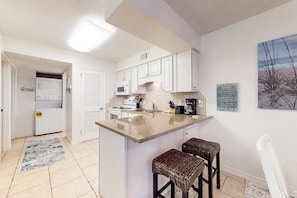 A kitchen with white cabinets, a microwave, a coffee maker, and a bar area with stools. - Beachfront condo with pool, hot tub, beach access & full kitchen (Gulf Shores)