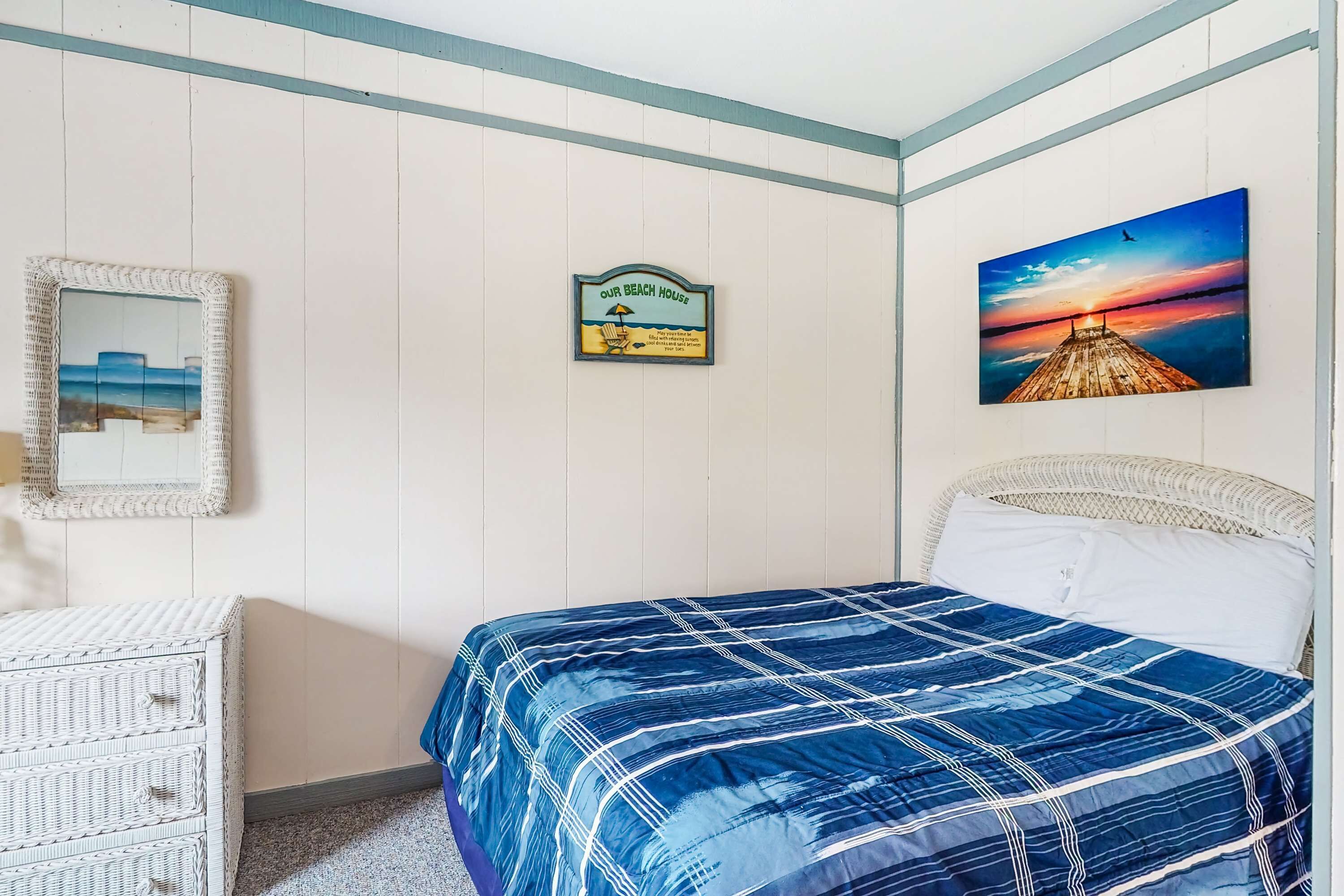 A bedroom with a bed, a wicker dresser, a framed picture, a beach-themed sign, and a sunset painting.