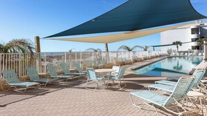 A poolside area with lounge chairs and a table, a pool, and a building in the background. - Gulf front beach condo with multiple community pools & sauna (Gulf Shores)