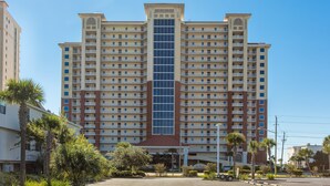 A multi-story hotel with a central glass facade, balconies, and a parking lot. - Gulf front beach condo with multiple community pools & sauna (Gulf Shores)