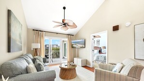 A spacious living room with a grey sofa, a striped armchair, a round coffee table, and a ceiling fan. A TV is mounted on the wall, and there is a view of a balcony with a pool. - Seabrook Exclusives | 2942 Atrium (Seabrook Island)