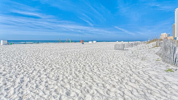 A sandy beach with a clear sky, a fence, and buildings in the background. - 5th-Floor Beach View Condo w/Balcony, Shared Pool, Outdoor Shower, A/C, WiFi (Gulf Shores)