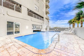 A swimming pool with a clear blue water, surrounded by lounge chairs and palm trees, adjacent to a building with balconies and windows. - Family-friendly Gulf-front condo with private balcony, hot tub, pool, & gym (Gulf Shores)