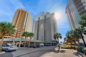 A multi-story hotel with a prominent logo on the facade, a parking area with several cars, and palm trees lining the street. - Family-friendly Gulf-front condo with private balcony, hot tub, pool, & gym (Gulf Shores)