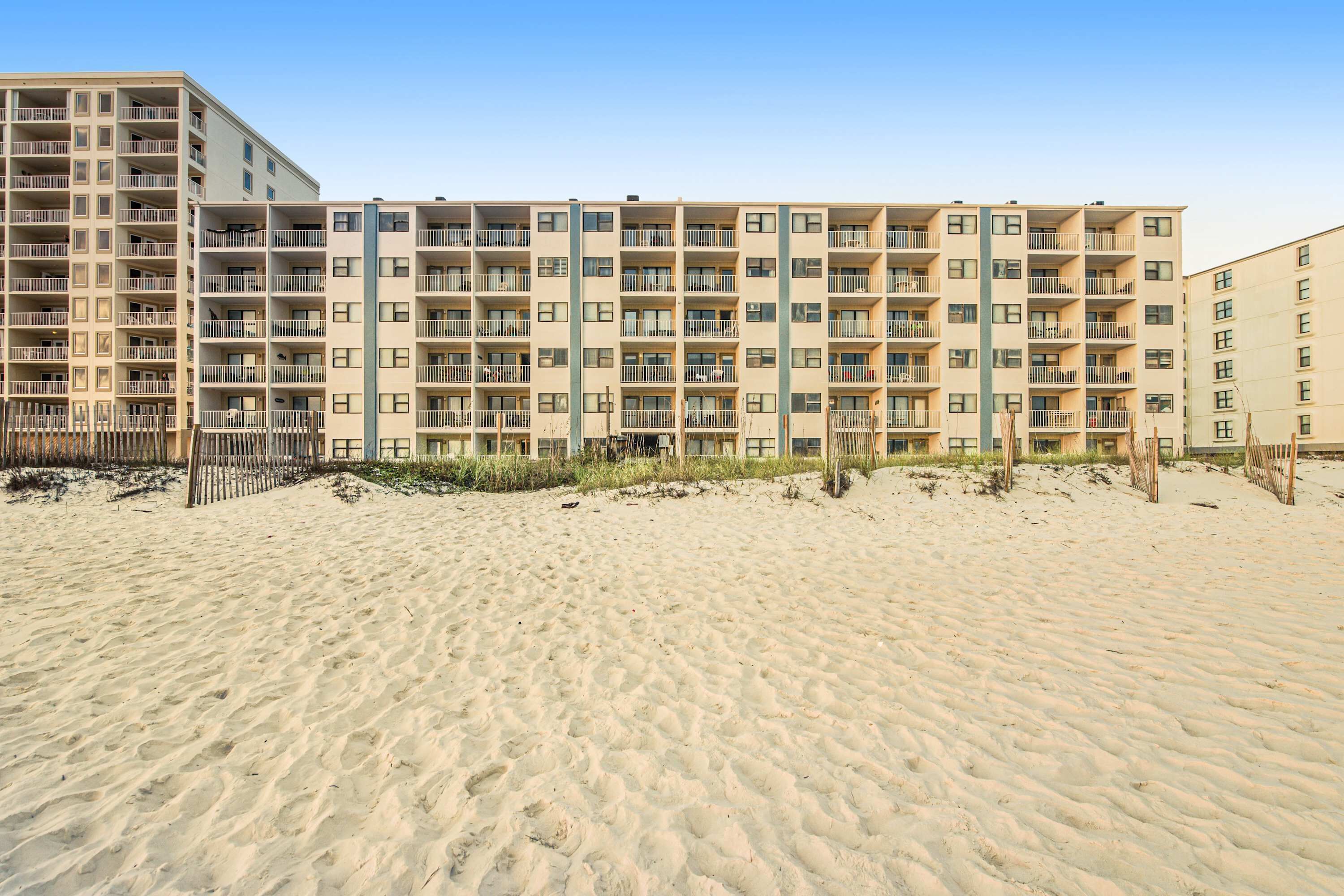 A multi-story hotel with balconies, a sandy beach in front, and a clear sky.