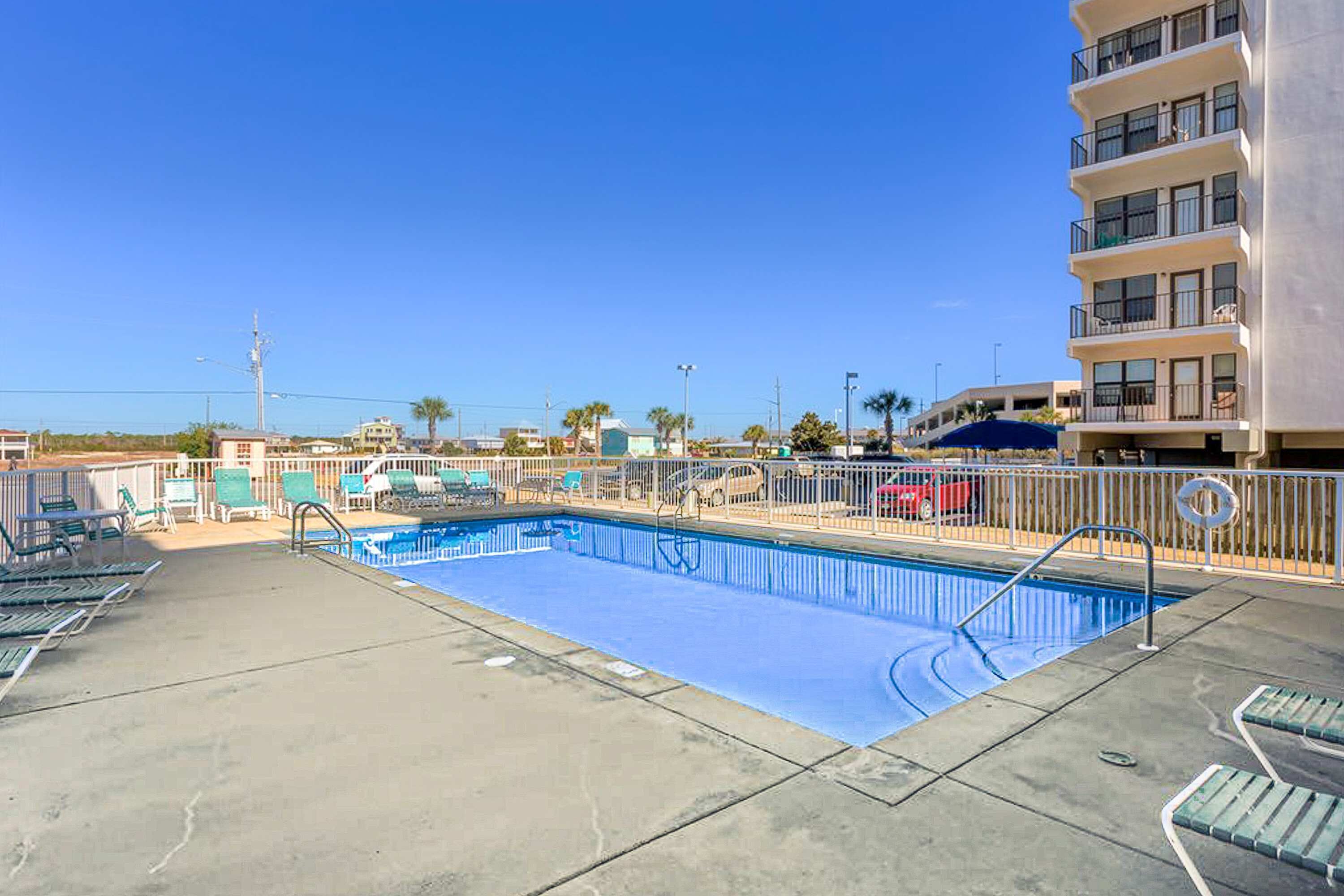 Une piscine avec des chaises longues, un espace délimité par une clôture et, au fond, un immeuble à plusieurs étages.