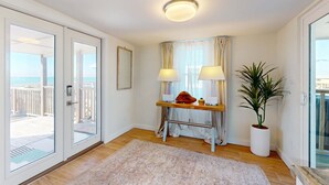Una habitación con piso de madera, una puerta blanca, una puerta de cristal que da a un balcón, una pequeña mesa de madera con dos lámparas, una planta en maceta y un lámpara en el techo.